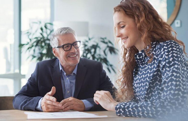 A man and a woman smiling and talking to each other while looking at papers on the desk in front of them.